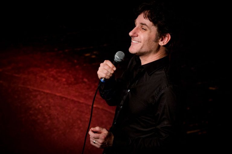 Tom Stade at the mic on a dark red stage