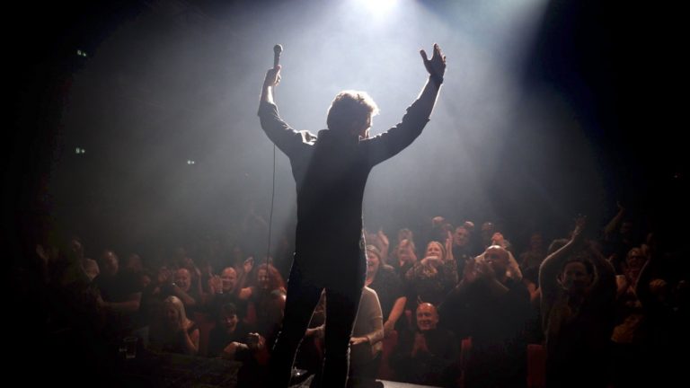 Tom Stade on stage, arms raised to a packed crowd