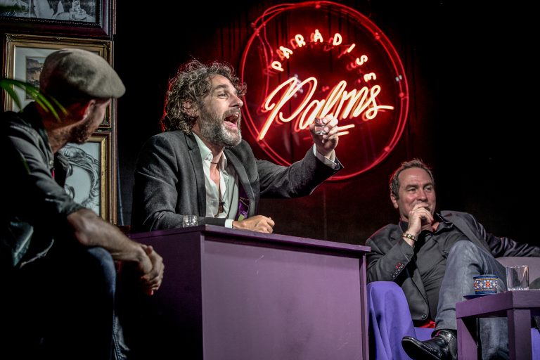 Tom Stade seated at a desk on stage beside a neon sign